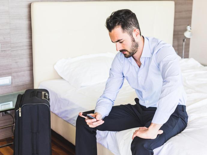 Uomo seduto su un letto con valigia, guarda il telefono.