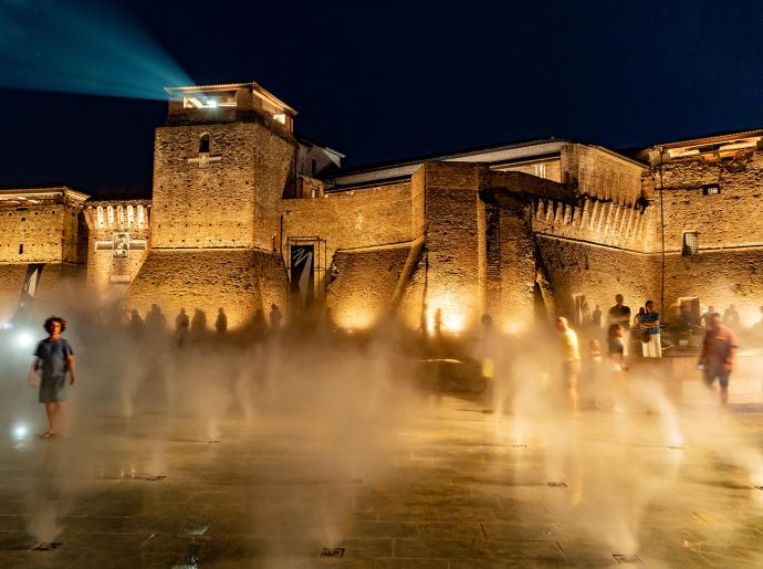 Castello illuminato di notte con nebbia e persone sul piazzale.