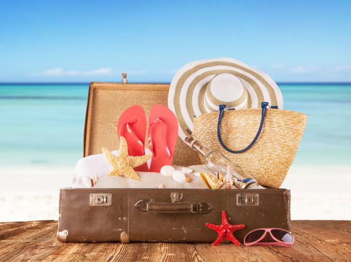 Offener Koffer am Strand mit Sommeraccessoires: Hut, Flip-Flops, Tasche und Muscheln.