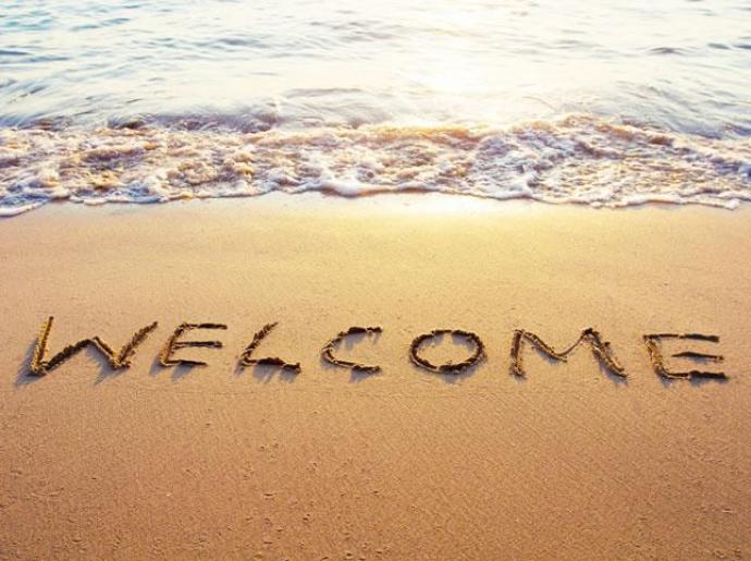 Parola 'Welcome' scritta sulla sabbia con onde sullo sfondo.
