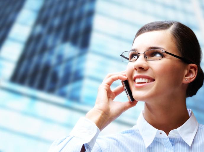Femme souriante au téléphone avec lunettes, fond de bâtiment moderne.